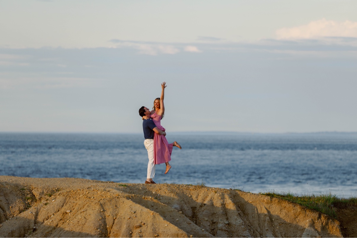 A newly engaged couple celebrating on a Long Island bluff above the ocean, the moment after she said yes