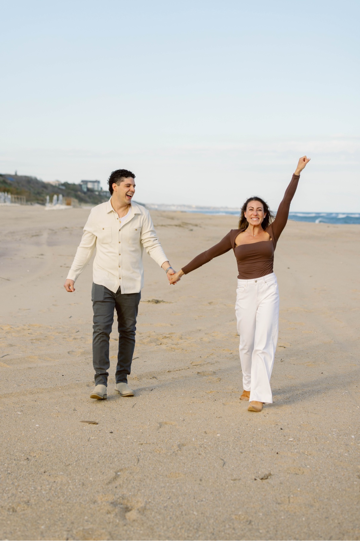 Couple celebrating their engagement on a wide Long Island beach, her arm raised in joy