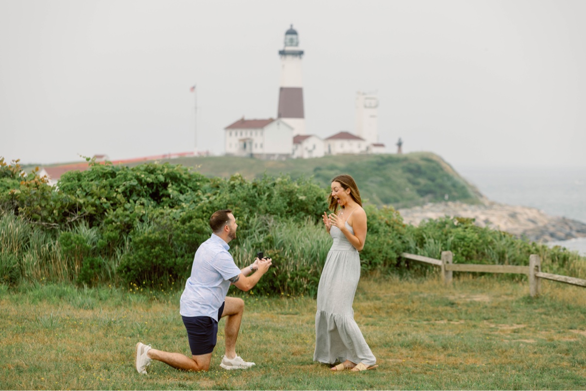 On one knee proposing in front of the Montauk Lighthouse, Long Island — her hands raised in surprise