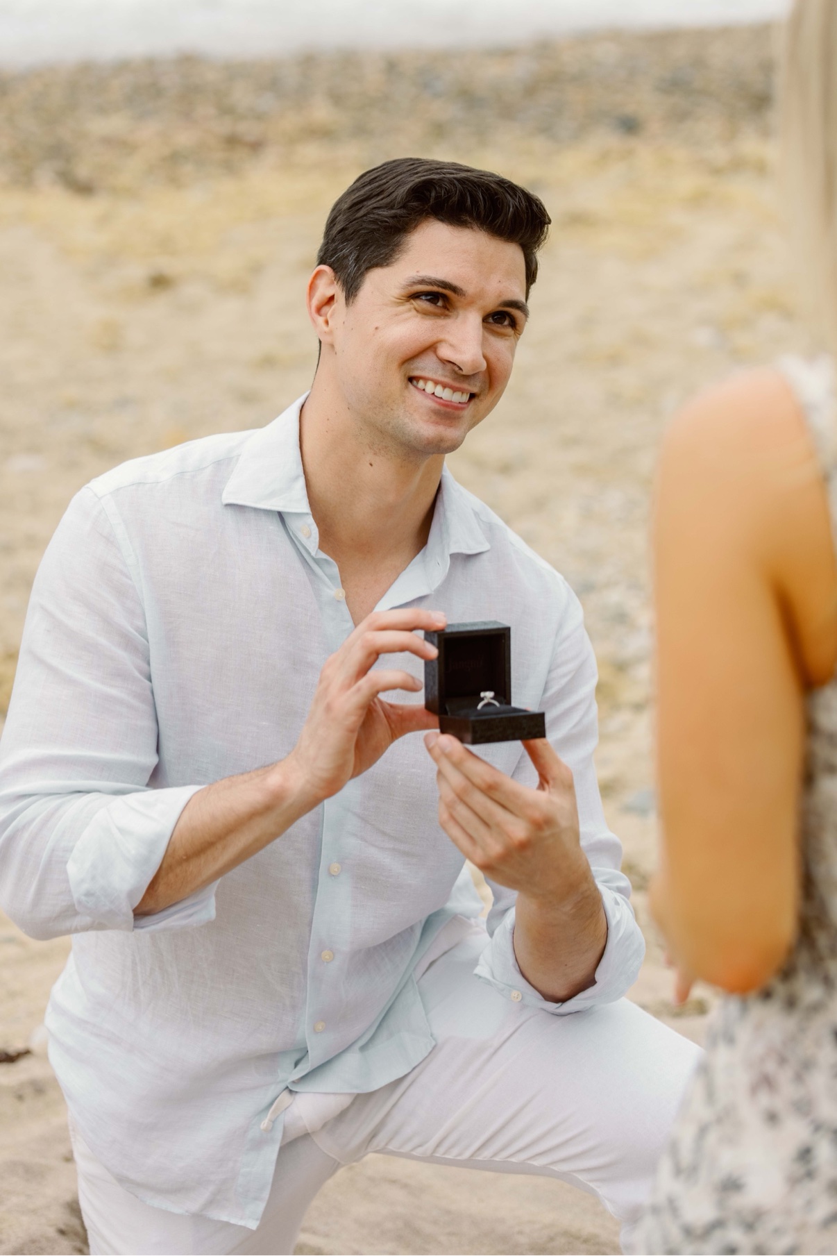 Surprise proposal at Montauk Lighthouse, Long Island — on one knee on the beach, holding the open ring box up to her with the bluffs behind them