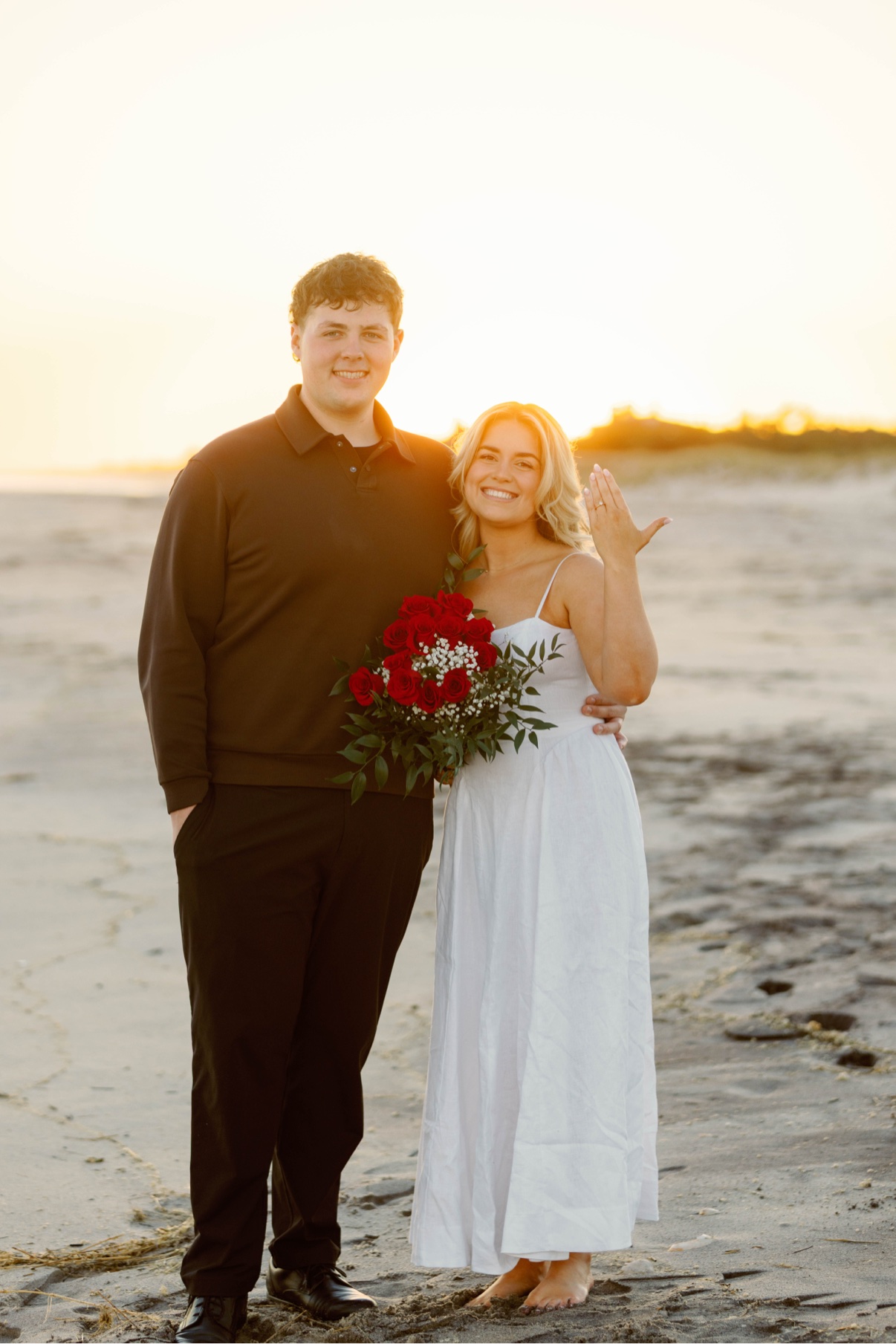 Surprise proposal at Cooper's Beach, Southampton, Long Island — a newly engaged couple at sunset, she raises her hand to show the ring, a bouquet of red roses in her arm