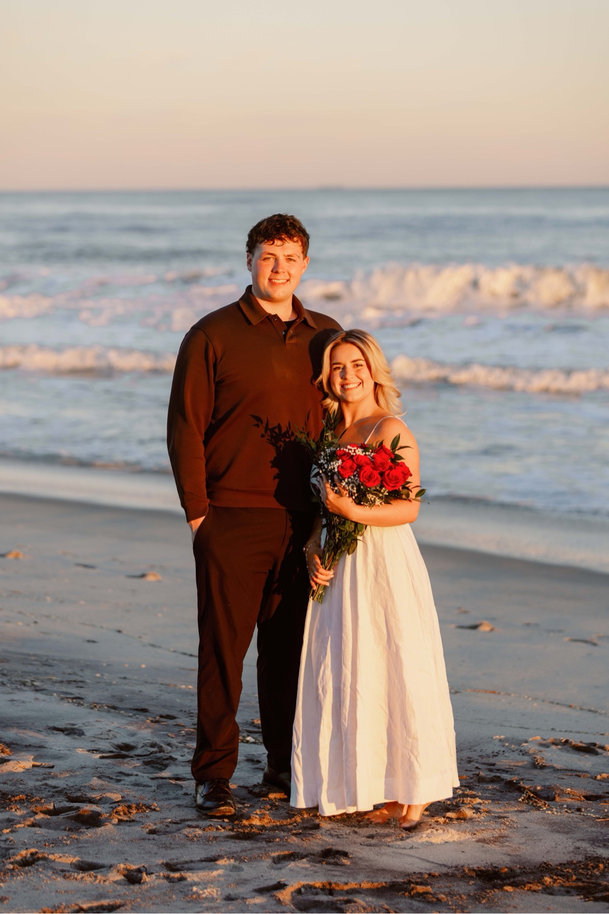 Newly engaged couple standing at the surf line on Cooper's Beach, Southampton, Long Island at sunset, her red rose bouquet in hand