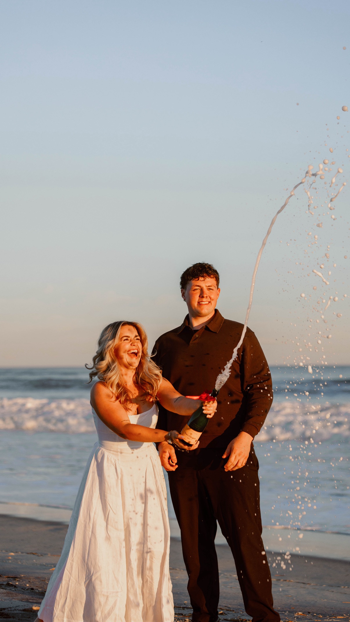 A couple popping champagne on Cooper's Beach, Southampton, Long Island to celebrate their engagement at sunset, both laughing