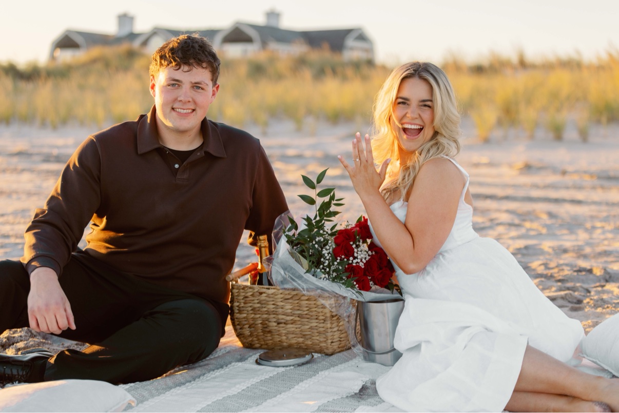 Engagement photos at Cooper's Beach, Southampton, Long Island — a couple seated on a picnic blanket on the sand, she shows her new ring, the beach houses behind them
