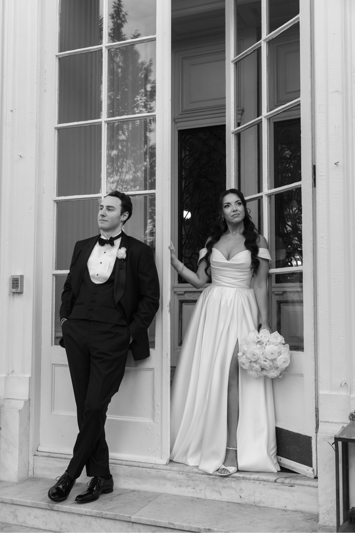 Groom in a tuxedo and bride in a white dress standing at tall French doors at Bourne Mansion