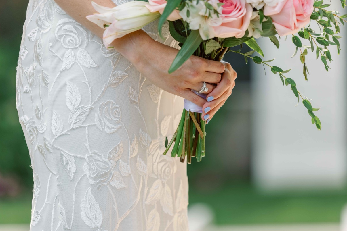 Close-up of the bride&rsquo;s hands holding her bouquet over her lace gown, her ring visible