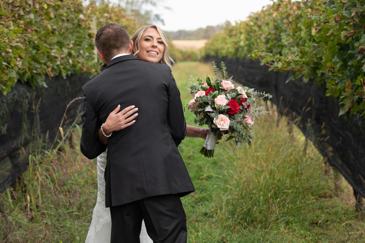 Bride smiling over the groom's shoulder as they embrace between vineyard rows